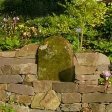 Trockenmauer aus Bergischer Grauwacke im Garten von Iris Ney. Den Grabstein aus Sandstein (Hundegrab) fand ich auf dem Gelände und habe ihn als Rückenlehne eines 'Thrones' in die den Hang abfangende Trockenmauer eingebaut.